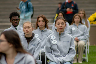 Remembrance Scholar Sasha Temerte, seated next to fellow scholar Karina Freeland, wears a pin with a small portrait of Karen Lee Hunt, the student Temerte represents. Temerte sits in seat 32K, one of the 35 chairs displayed outside of Hendricks Chapel. 